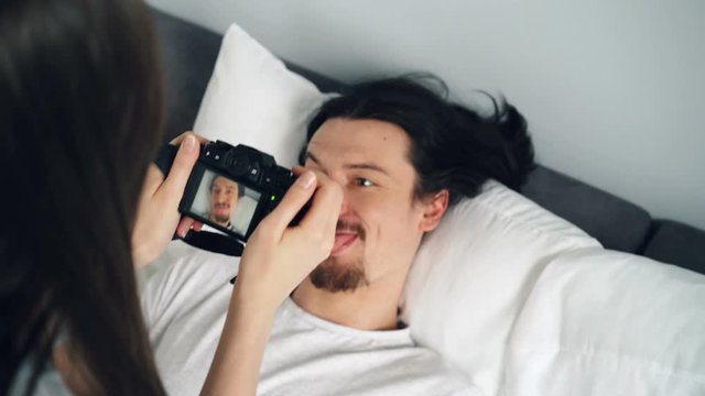 Playful young man is posing with funny face at home in bed while girl is using modern professional camera taking pictures. Photography, youth and hobby concept.