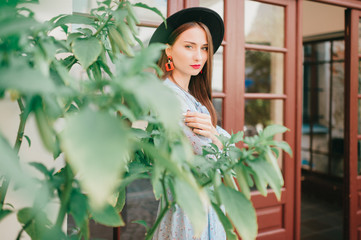 Romantic girl in blue dress and elegant hat posing in french style yard with garden and green bushes