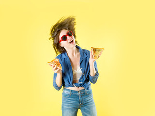 Fashionable girl on a yellow background in a blue shirt, in red sunglasses and with flying hair holds a piece of pizza in her hands