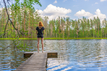Fototapeta na wymiar Cute ginger girl standing on wooden planked footway in summer day against beautiful landscape of northern lake and forest. Travelling and discovering distant places of Earth. Karelia, Russia