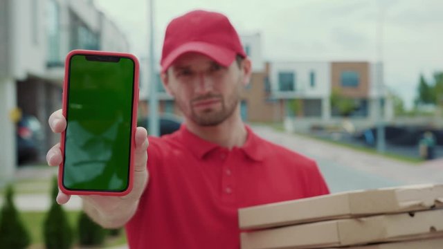 Portrait of serious pizza courier showing green smartphone mock up and holding delivery boxes. Close-up of young delivery man in red uniform with paypass mobile phone payment delivering order staying