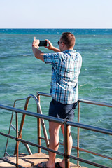 Tourist and traveler on beach pier is taking pictures using camera his smart phone of sea coast, environment and nature landscape. Man with sunglasses on vacation in hotel resort on beach platform.