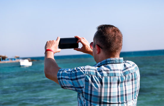 Tourist and traveler on beach pier is taking pictures using camera his smart phone of sea coast, environment and nature landscape. Man with sunglasses on vacation in hotel resort on beach platform.