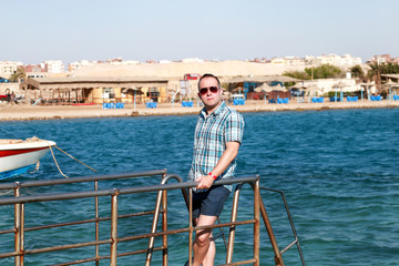 Portrait of tourist and traveler is posing and walking on beach pier, enjoys in sun and view of sea landscape. Man with sunglasses on vacation in summertime in hotel resort on sea jetty platform.