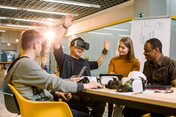 Teamwork trying virtual reality glasses for work in creative office. Young Asian man using new technology vr goggles while his coworkers talking sitting at the table