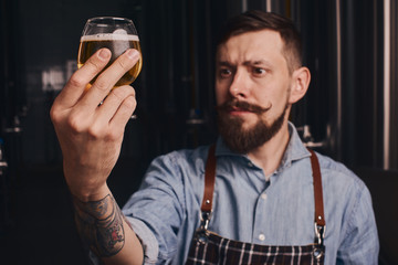 Bartender in the apron checks beer quality holding it in front of himself.