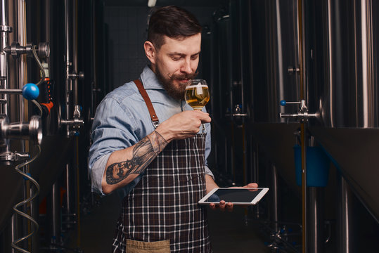 Tattooed man tastes beer from the glass he holds.