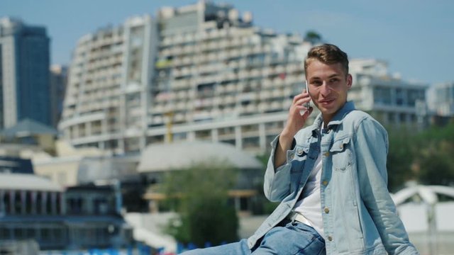 smiling young man in fashionable clothes sits outside and talks on phone against large hotel close view slow motion