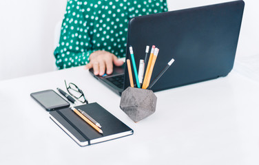 woman in green blouse typing on keyboard laptop, concrete holder with pencils and pens, notebook, smartphone, glasses and black paperweight on white table, education office concept background.