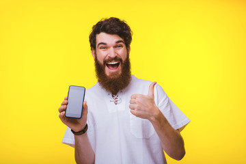 Exited bearded man is holding a smartphone and showing thumb up near yellow wall.
