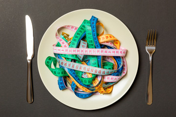 Top view of colorful measuring tapes on plate in the form of spaghetti with knife and fork on black...