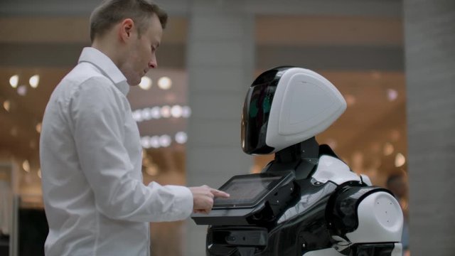 A man in a shirt communicates with a white robot asking questions and pressing the screen with his fingers.