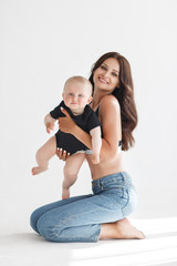 Young woman mother in denim overalls holds a baby child in her arms. White background in the studio.Mother with baby isolated on white. Mothers Day, love family, parenthood childhood concept. Happy fa