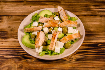Fresh salad with chicken meat, feta cheese, avocado, green olives and lettuce leaves in ceramic plate on wooden table
