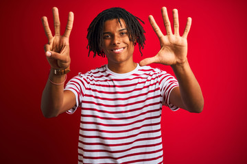 Young afro man with dreadlocks wearing striped t-shirt standing over isolated red background showing and pointing up with fingers number eight while smiling confident and happy.