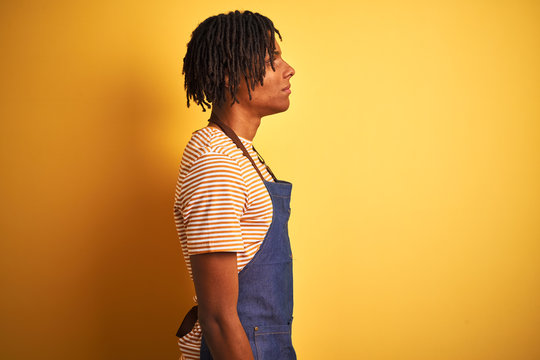 Afro american barista man with dreadlocks wearing apron over isolated yellow background looking to side, relax profile pose with natural face with confident smile. - Powered by Adobe