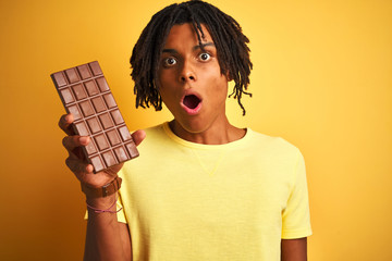Afro american man with dreadlocks eating chocolate bar over isolated yellow background scared in...