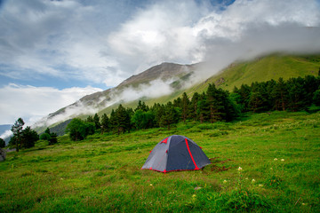 tourist tent stands on a green meadow against the backdrop of mountain slopes and peaks with clouds and blue sky