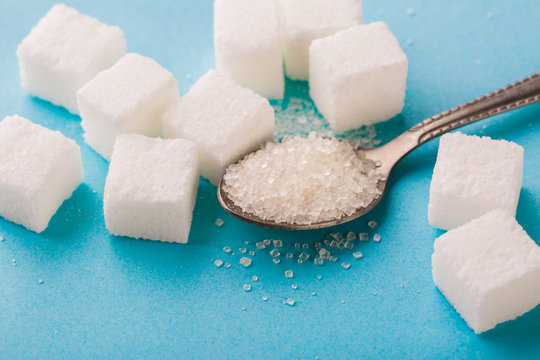 Granulated sugar in spoon with cube on blue background.