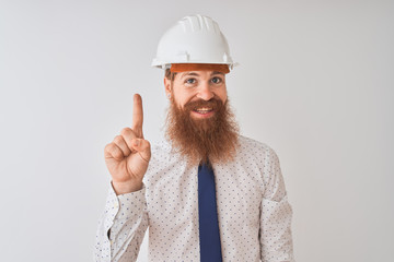 Young redhead irish architect man wearing security helmet over isolated white background showing and pointing up with finger number one while smiling confident and happy.