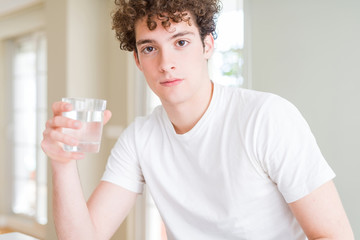 Young man drinking a glass of water at home with a confident expression on smart face thinking serious