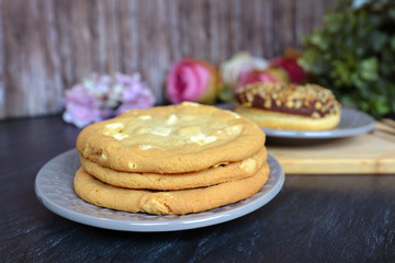 Three delicious looking home baked white chocolate chips cookies arranged on a plate with blurry pastry in background