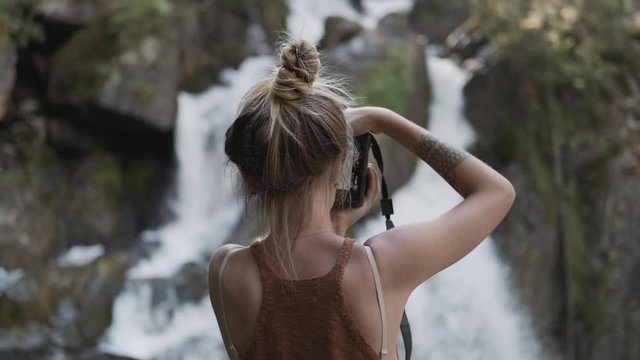 A young paparazzi girl takes pictures of a beautiful large waterfall standing on stones in the jungle. Back view