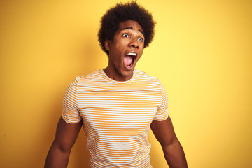 American man with afro hair wearing striped t-shirt standing over isolated yellow background angry and mad screaming frustrated and furious, shouting with anger. Rage and aggressive concept.