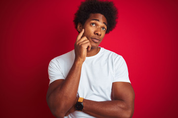 Fototapeta na wymiar Young american man with afro hair wearing white t-shirt standing over isolated red background with hand on chin thinking about question, pensive expression. Smiling with thoughtful face.