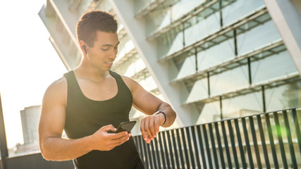 Cheerful mixed race athlete looking at his smart watch on the wrist with mobile phone in other hand