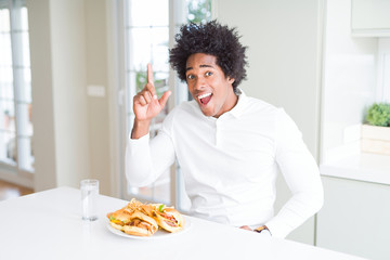 Fototapeta na wymiar African American hungry man eating hamburger for lunch pointing finger up with successful idea. Exited and happy. Number one.