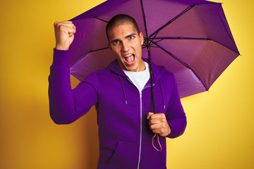 Young handsome man wearing purple umbrella over yellow isolated background annoyed and frustrated shouting with anger, crazy and yelling with raised hand, anger concept