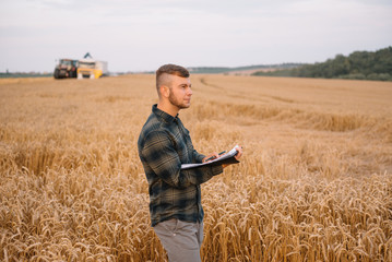 Naklejka na ściany i meble Young agronomist man standing on wheat field checking quality while combine harvester working.