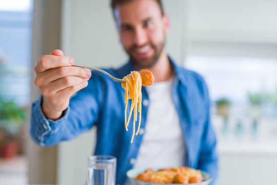 Handsome man eating pasta with meatballs and tomato sauce at home while smiling at the camera