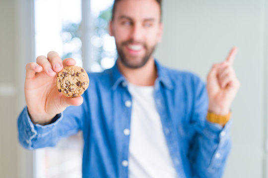 Handsome man eating chocolate chips cookies very happy pointing with hand and finger to the side