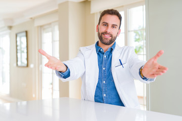 Naklejka na ściany i meble Handsome doctor man wearing medical coat at the clinic looking at the camera smiling with open arms for hug. Cheerful expression embracing happiness.