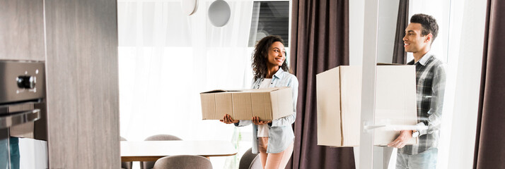 panoramic shot of african american husband and wife holding boxes and looking at each other while walking into house