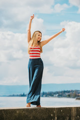 Outdoor fashion portrait of beautiful young woman posing by the lake, wearing red and white stripe vest and denim wide leg jean