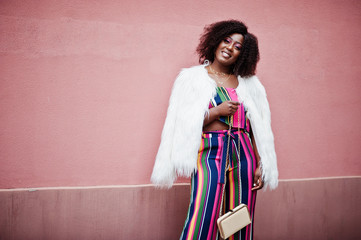 Fashionable african american woman in striped jumpsuit with fluffy faux fur coat and handbag posed...