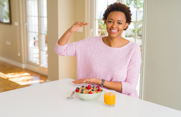 Young african american woman having healthy breakfast in the morning at home gesturing with hands showing big and large size sign, measure symbol. Smiling looking at the camera. Measuring concept.
