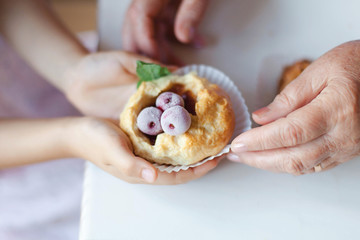 Kid gives homemade cupcake to senior woman. Hands of child girl and retired grandmother. Baked pastries with berries and mint leaves. Buns with powdered sugar. Family is cooking pies at home kitchen.