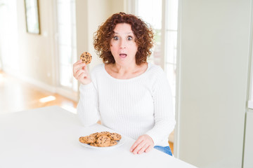 Senior woman eating chocolate chips cookies at home scared in shock with a surprise face, afraid and excited with fear expression