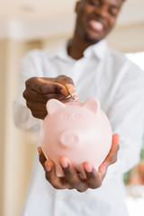 Handsome young african man putting a coin inside piggy bank smiling, excited about investment