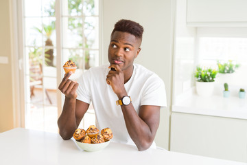 African american man eating chocolate chips muffin serious face thinking about question, very confused idea