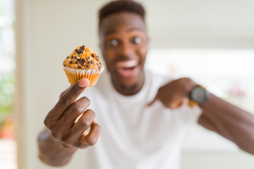 African american man eating chocolate chips muffin with surprise face pointing finger to himself