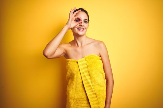 Young beautiful woman wearing towel after shower over yellow isolated background doing ok gesture with hand smiling, eye looking through fingers with happy face.