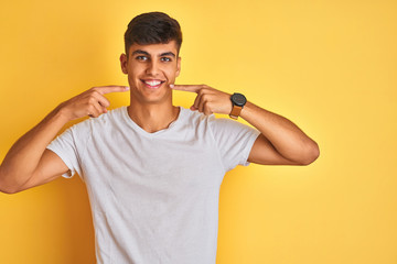 Young indian man wearing white t-shirt standing over isolated yellow background smiling cheerful showing and pointing with fingers teeth and mouth. Dental health concept.