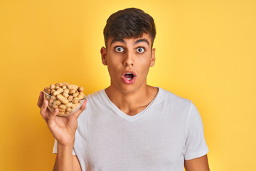 Young indian man holding bowl with peanuts over isolated yellow background scared in shock with a...