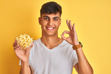 Young indian man holding bowl with extruded corn standing over isolated yellow background doing ok sign with fingers, excellent symbol