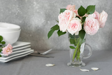 Tea roses in a glass on a background of a stack of white dishes on a table with a gray linen tablecloth.